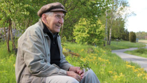Ein alter Mann sitzt lächelnd an einem Wegrand inmitten blühender Felder.