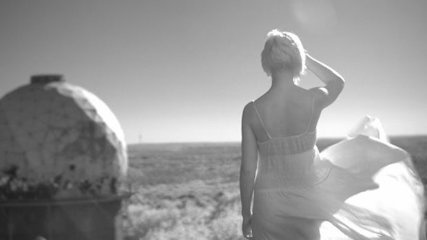 Eine junge Frau blickt mit dem Rücken zur Kamera über eine weite Landschaft in grellem Sonnenlicht.